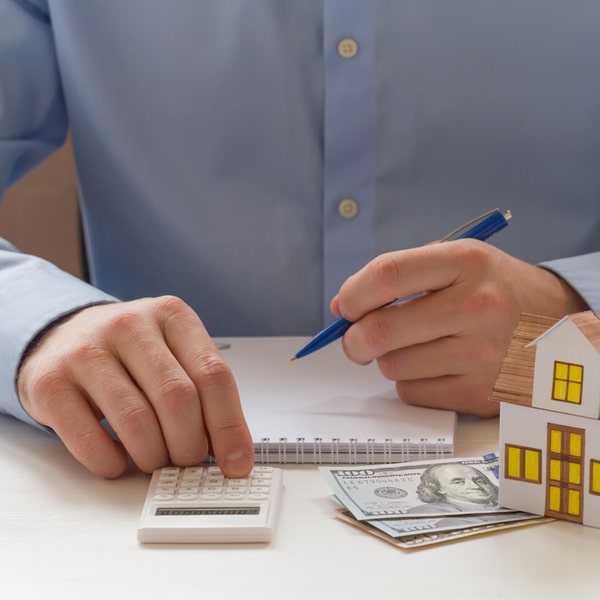 Person doing calculations with a pen, calculator, and money next to a model house, highlighting the benefits of a high credit score in financial planning and real estate investments.